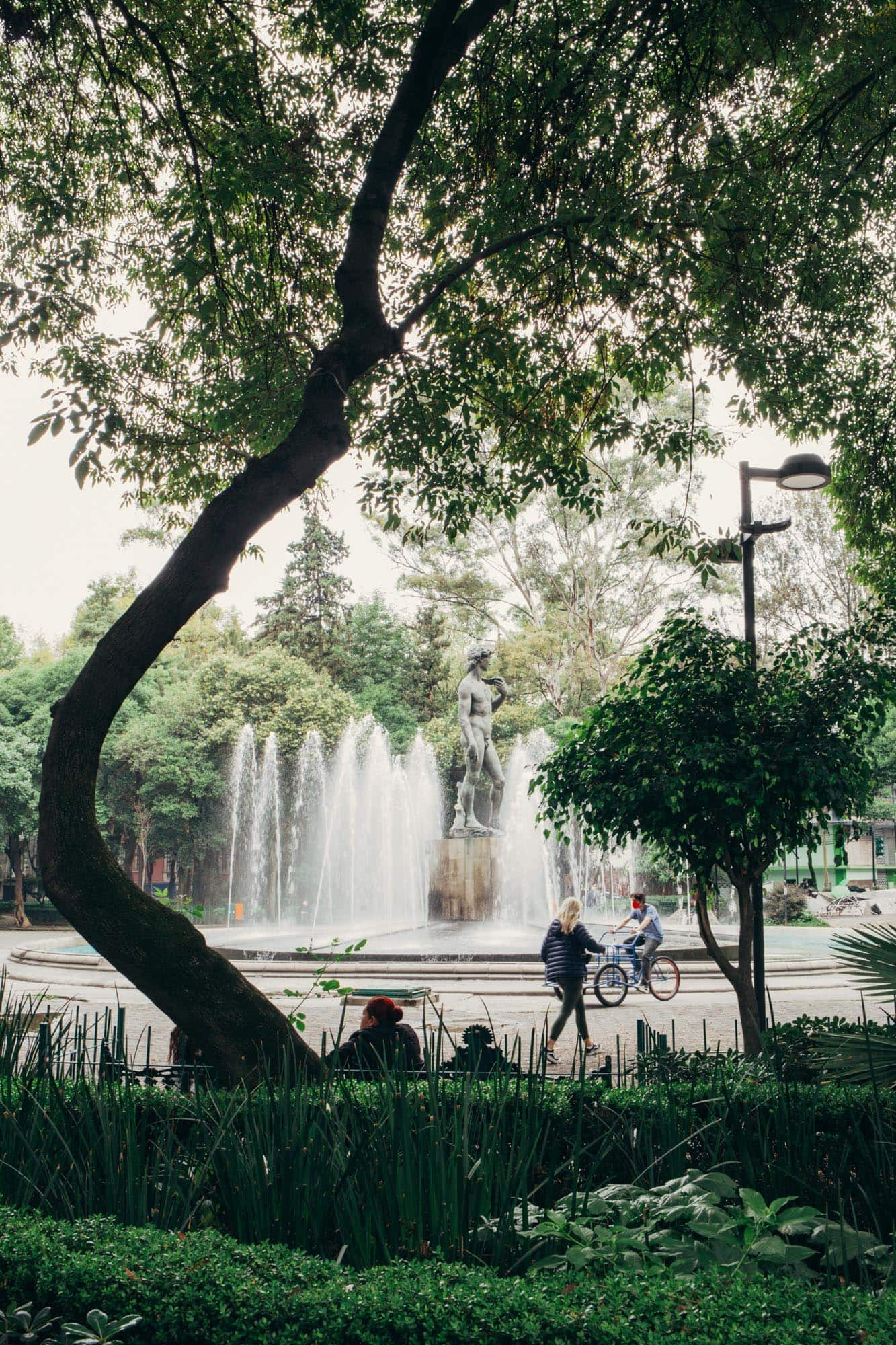 A serene park scene with a large fountain at its center, featuring a statue of a man. The fountain is surrounded by tall, arching trees that provide ample shade. People can be seen enjoying the park, with one person cycling and another walking nearby, highlighting the park
