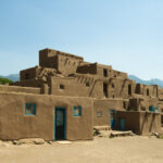 Adobe-style buildings with blue doors, set against a clear sky and mountains in the background.