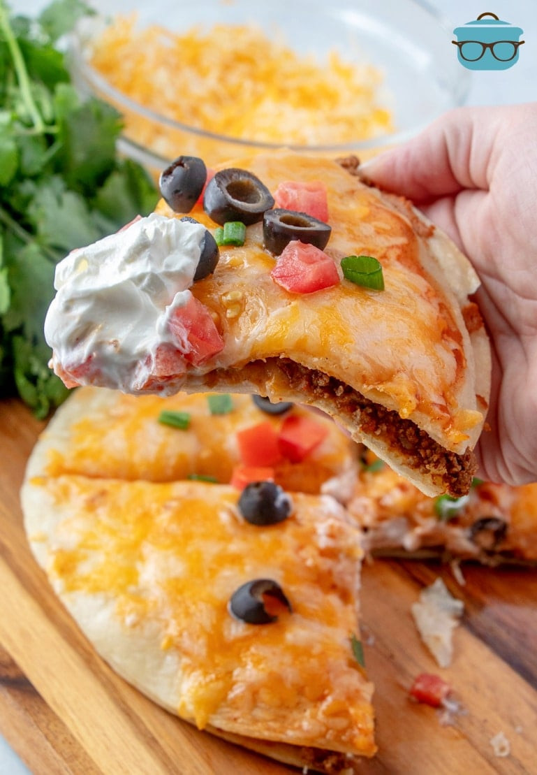 A close-up of a slice of Mexican pizza being held, showing the crispy tortilla and the layers of toppings.