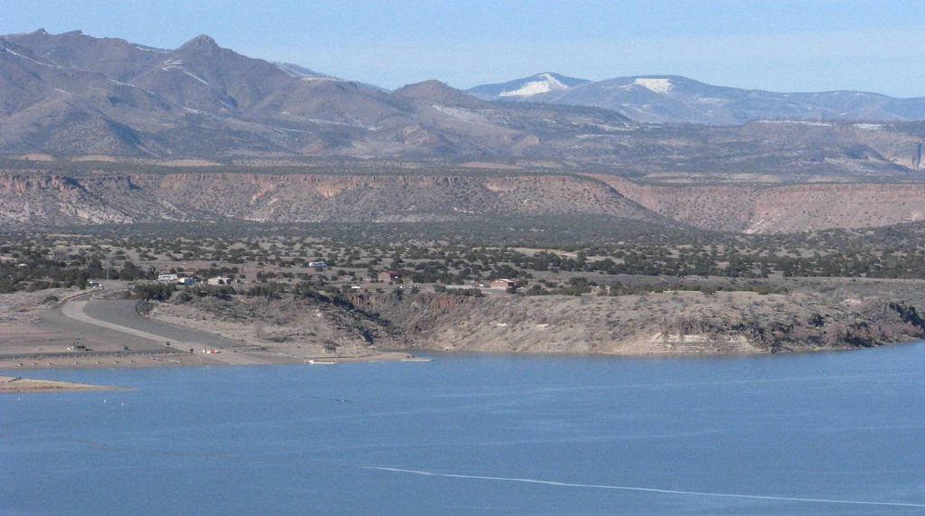 A picturesque lake surrounded by mountains beneath a clear blue sky, with a winding road tracing the shoreline at Cochiti Lake, New Mexico.