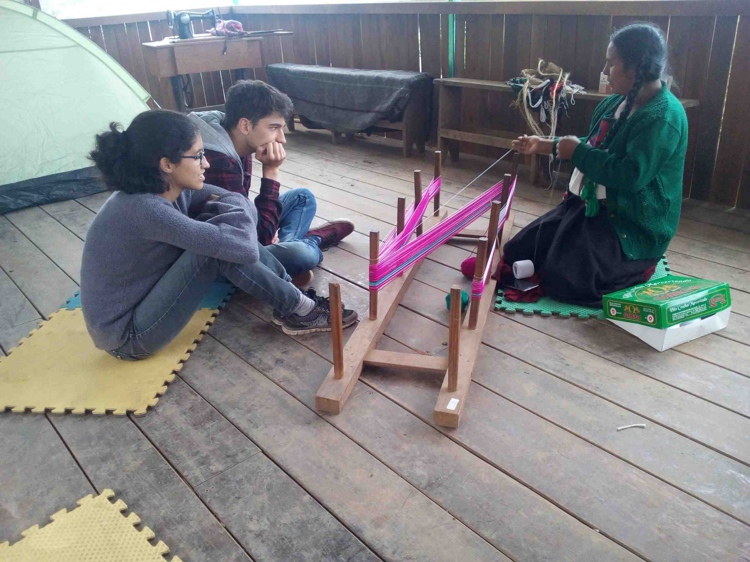 A community woman teaches handicraft weaving to two travelers, showcasing cultural immersion in Mexico.