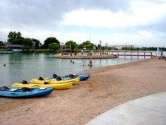 Vibrant kayaks resting on a sandy shore beside a calm waterway, with people swimming and a small island dotting the backdrop, Lake Carlsbad Recreation Area, New Mexico.