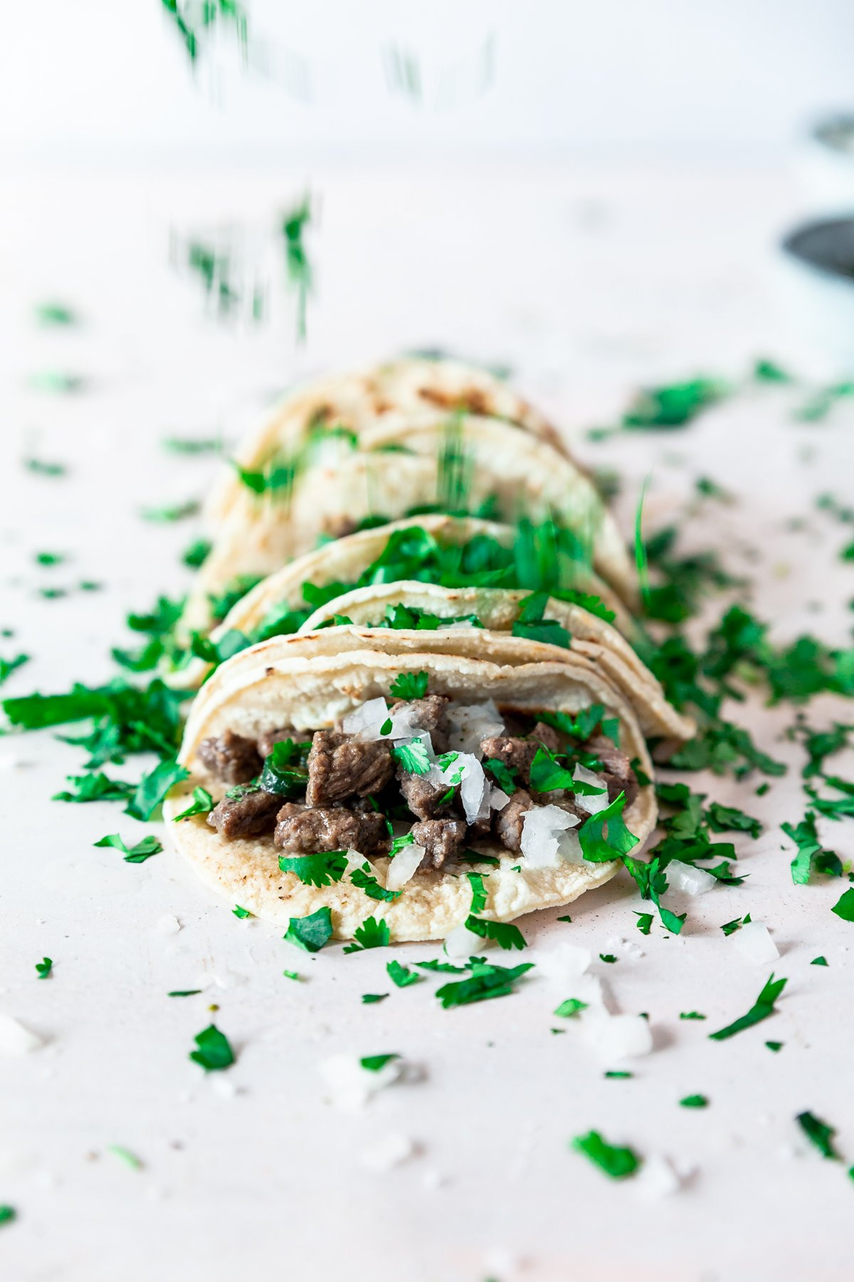 straight ahead view of five carne asada mexican street tacos being sprinkled with fresh cilantro. the easy mexican recipe is topped with fresh onion as well, and two light blue bowls are barely visible towards to the top-right of the image.