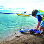 A child in a life jacket plays with toy boats and a truck on the shore of a lake, with a boat visible in the distance, Elephant Butte Lake State Park, New Mexico.