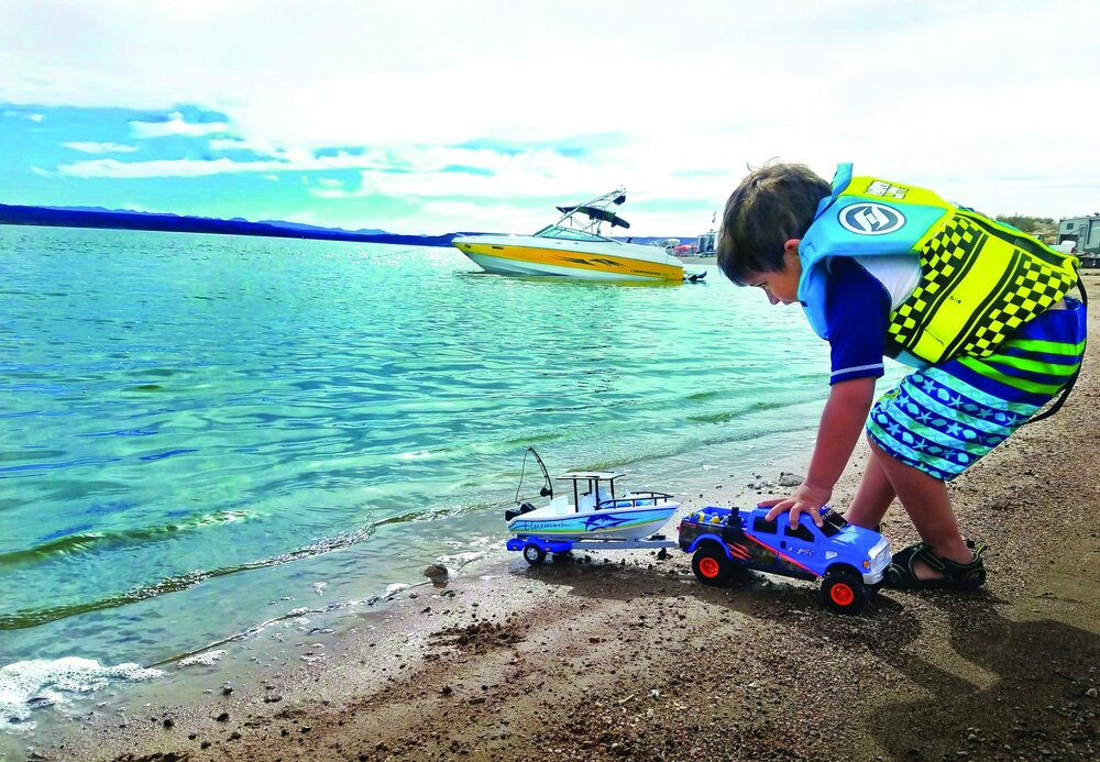 A child in a life jacket plays with toy boats and a truck on the shore of a lake, with a boat visible in the distance, Elephant Butte Lake State Park, New Mexico.