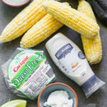 Elote ingredients including corn, mayonnaise, Mexican crema, cotija cheese, chili powder, cilantro, and lime.