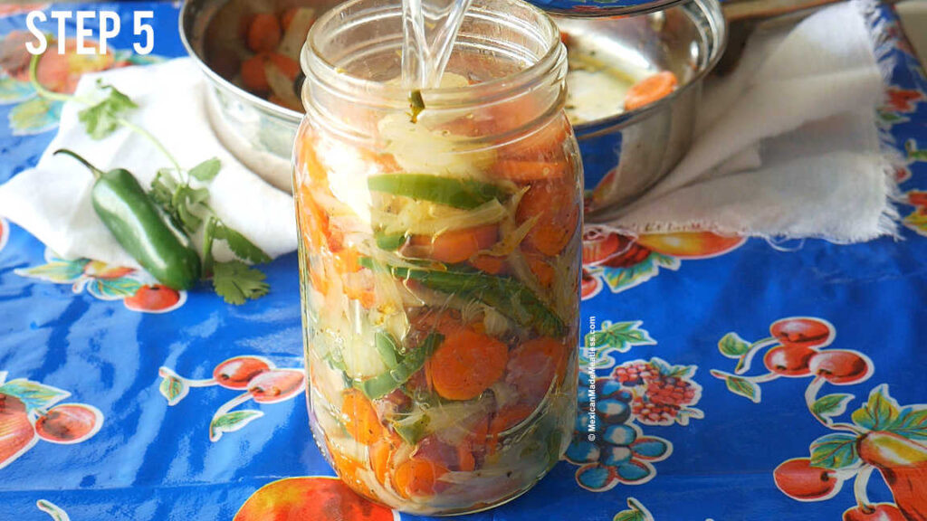 Filling a large mason jar with carrots, spices, and pickling brine, showcasing the layering of ingredients and the final step in preparing Mexican Spicy Pickled Carrots.