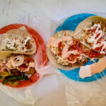 Fresh battered fish tacos topped with onions, coleslaw and spicy sauces. The plates are bright red and blue and are wrapped in see-through plastic.