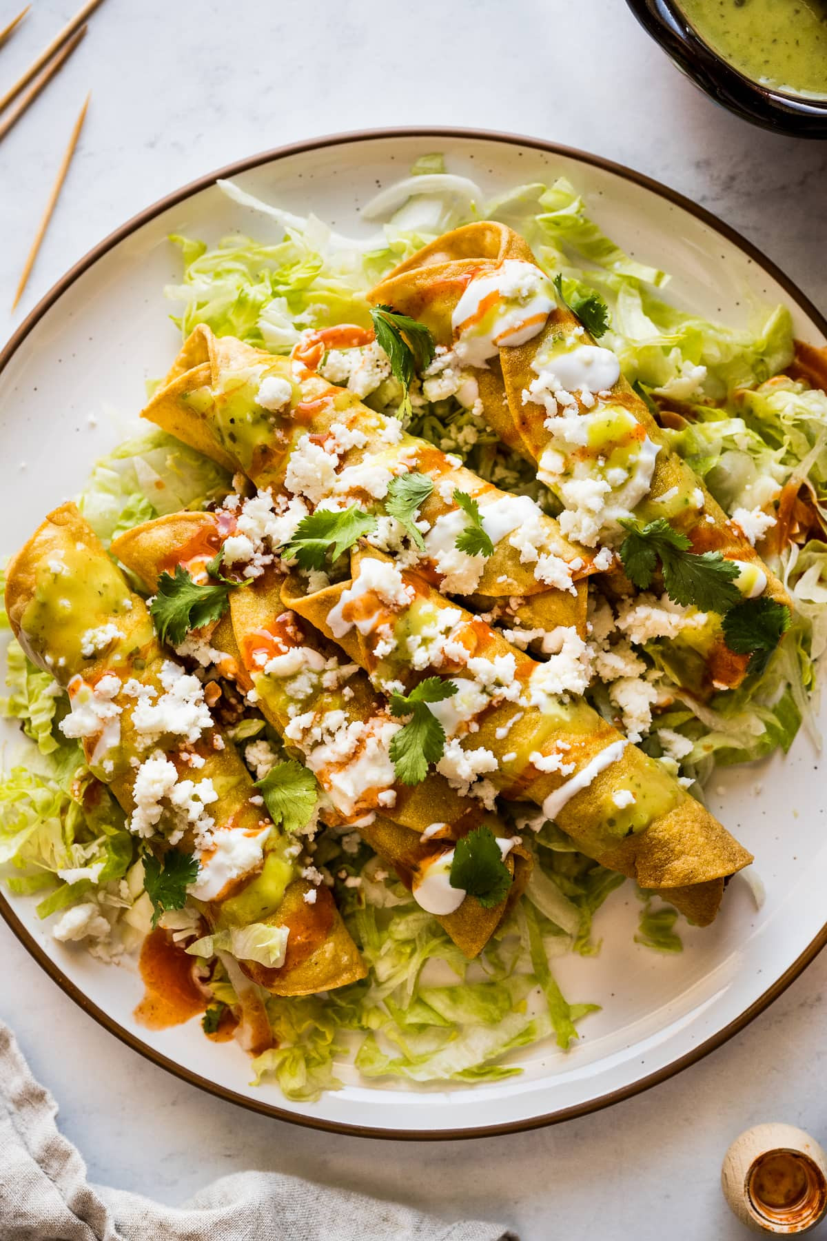 A plate of crispy chicken flautas topped with crema, hot sauce, salsa verde, lettuce, cilantro, and cotija cheese
