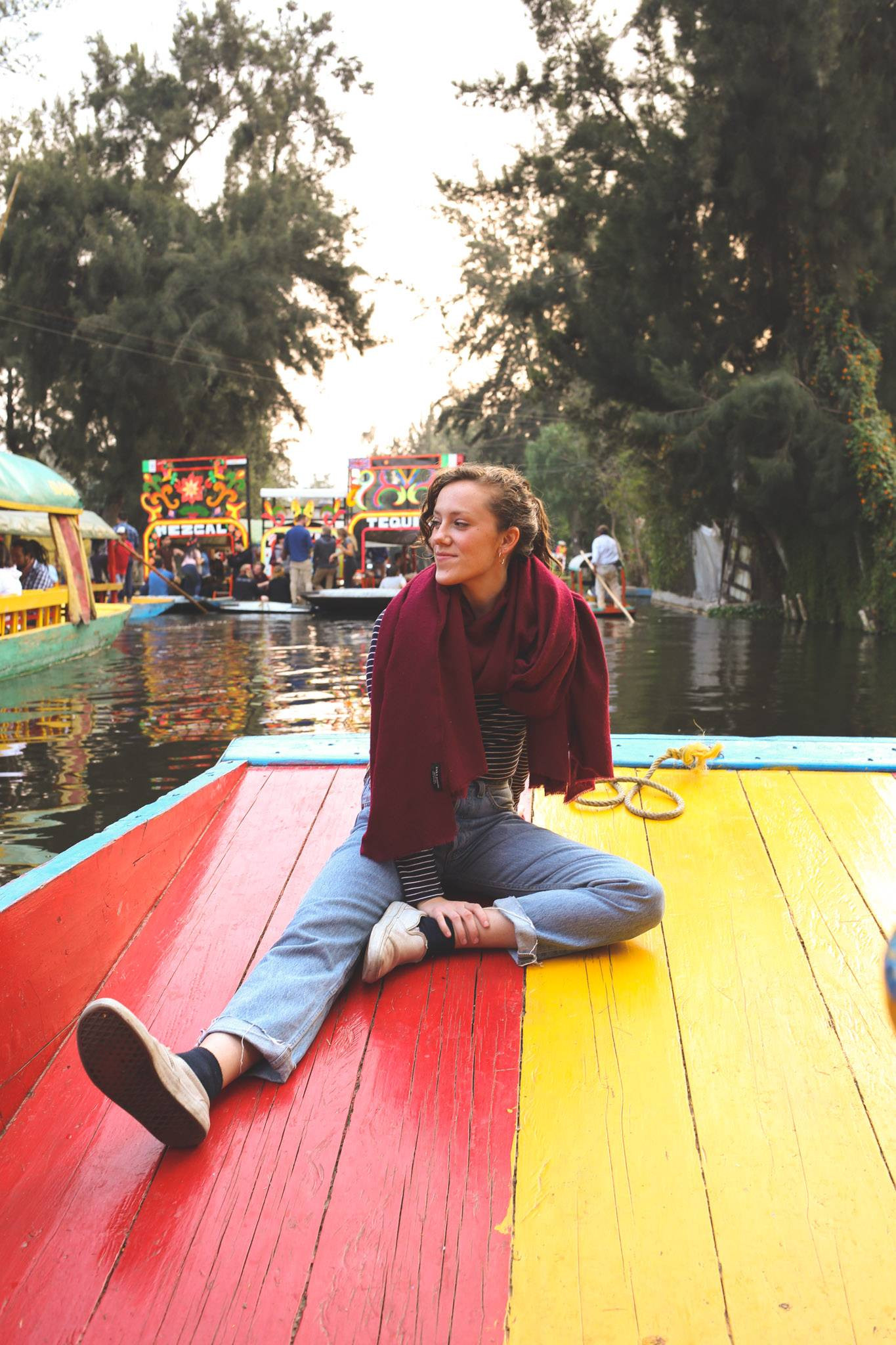 The image features a young woman sitting on a brightly colored boat with red and yellow stripes, enjoying a serene moment. She is surrounded by water and other decorated boats, suggesting a vibrant and lively atmosphere, likely in the Xochimilco canals in Mexico City. The background includes lush greenery and other people engaging in similar boat activities.