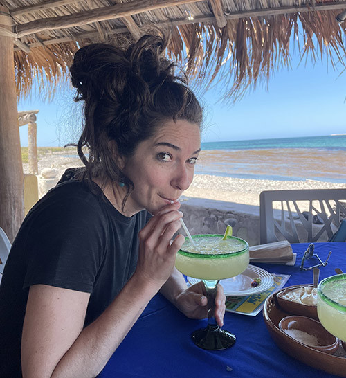 A woman sipping a large margarita at an outdoor table under a thatched roof, with a view of the beach and ocean in the background, Picazón providing the best margaritas in town with stunning beach views.