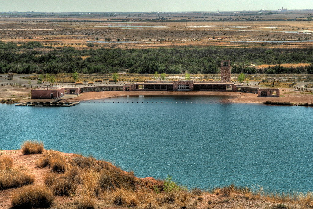 A tranquil blue lake surrounded by arid terrain, with a rustic building nestled along the shore at Lea Lake in Bottomless Lakes State Park, New Mexico.