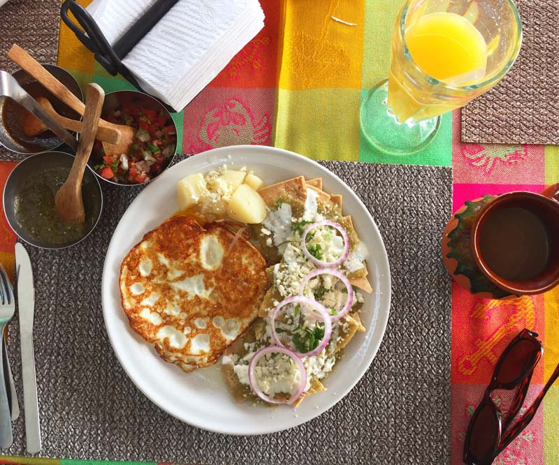 A breakfast plate featuring chilaquiles topped with cheese, red onion rings, and cilantro, served alongside a fried egg and a portion of boiled potatoes. The table setting includes condiments like salsa and pico de gallo, with a glass of orange juice and a cup of coffee on a colorful tablecloth, Los Mandiles offering traditional chilaquiles on the malecón for a great breakfast in Loreto.
