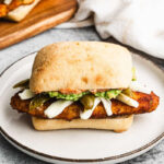 A Mexican Torta recipe with breaded, crispy chicken (chicken milanese), cheese, refried beans, guacamole, and pickled peppers on a white plate.