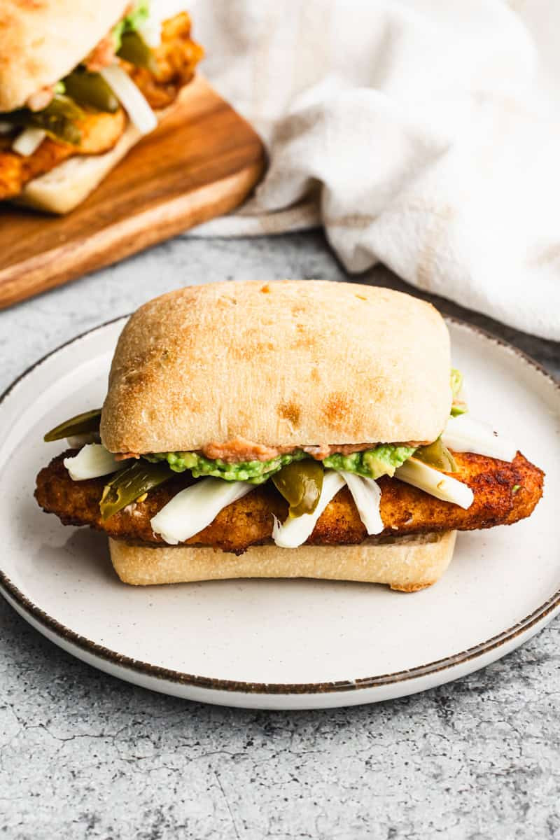 A Mexican Torta recipe with breaded, crispy chicken (chicken milanese), cheese, refried beans, guacamole, and pickled peppers on a white plate.