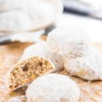 Mexican wedding cookies on a brown paper, with one cookie half eaten, and a plate of cookies in the background.