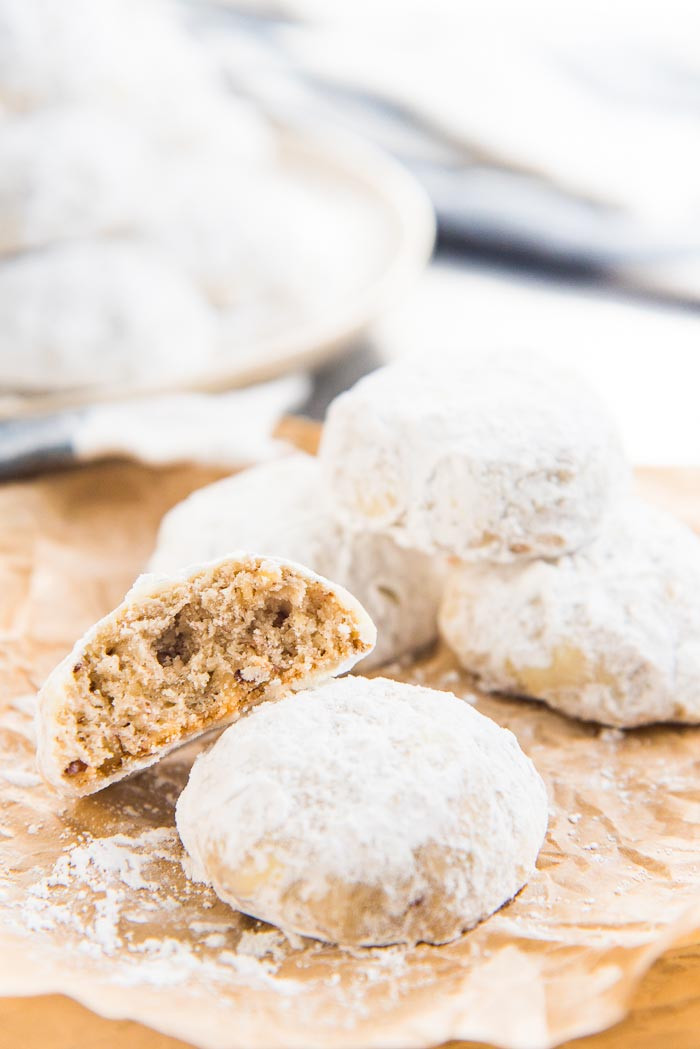 Mexican wedding cookies on a brown paper, with one cookie half eaten, and a plate of cookies in the background.