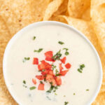 A bowl of white cheese dip topped with tomatoes and cilantro with tortilla chips next to the bowl