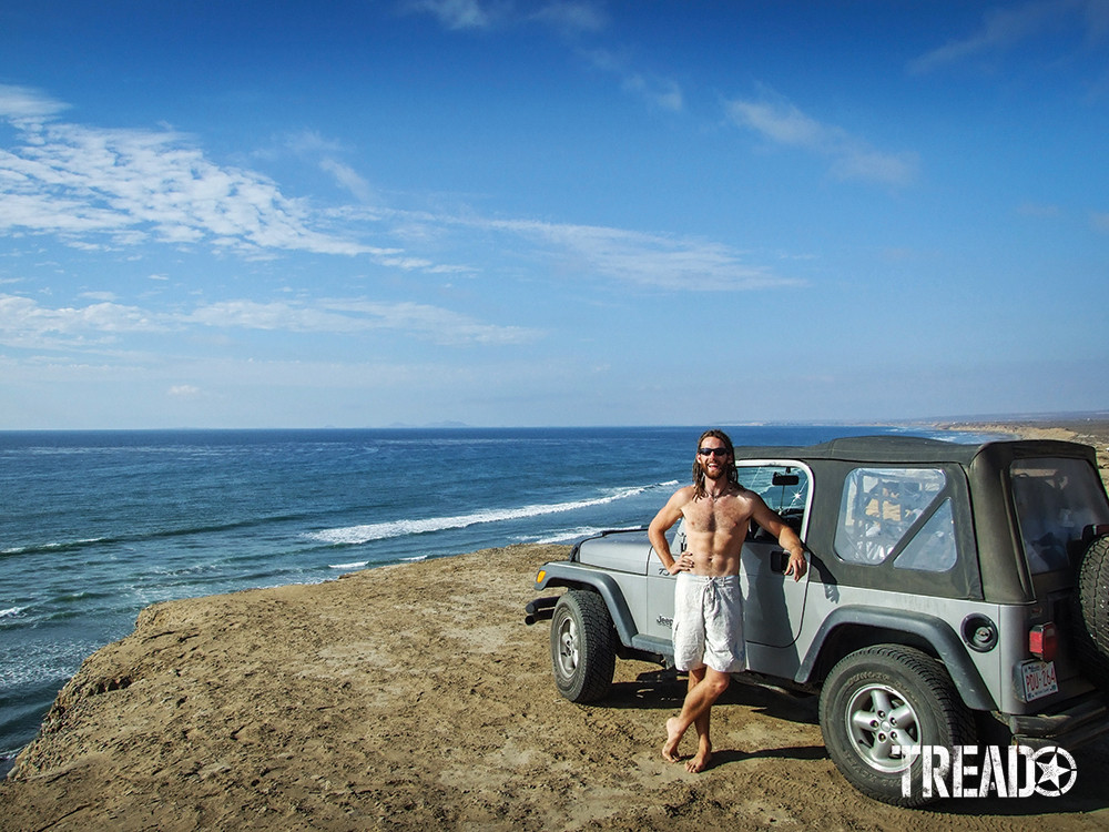Man and silver jeep next to ocean.