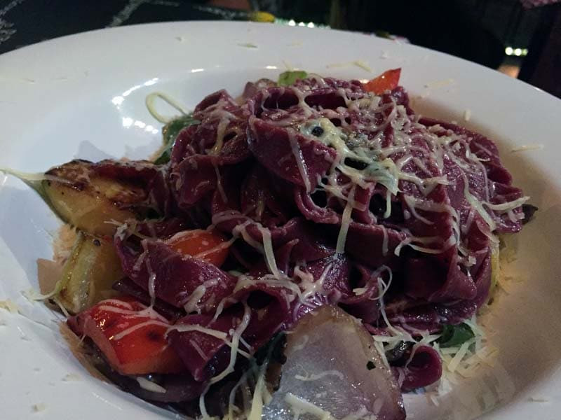 A plate of purple-hued pasta topped with shredded cheese, accompanied by grilled vegetables such as zucchini and red bell peppers, Mezzaluna offering unique purple fettuccini with fresh veggies.