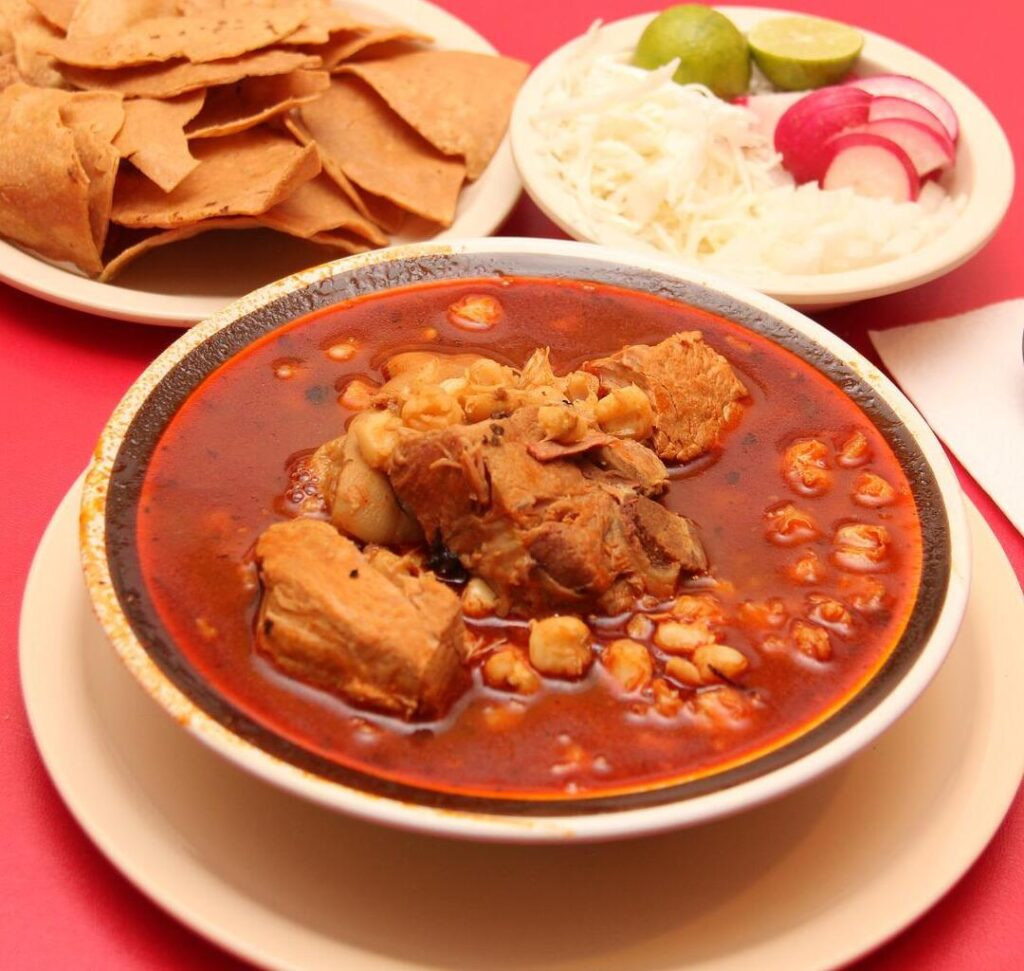 A bowl of red pozole with various toppings like shredded cabbage, lime wedges, and radishes