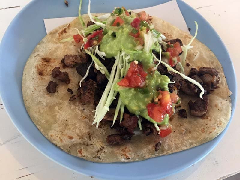 A taco on a flour tortilla filled with grilled beef, topped with shredded cabbage, guacamole, and diced tomatoes. The taco is served on a blue plate, El Rey del Taco presenting a fresh and colorful carne asada taco.