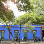 People line up in front of a one-story building painted blue on a city street shaded by trees.