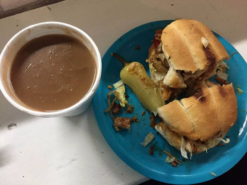 A plate with a sandwich made of toasted bread filled with shredded meat and lettuce, accompanied by a grilled pepper. Next to the plate is a cup of brown soup or broth, Torta and champurrado from La Super Torta offering a taste of authentic Mexican street food.