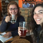 Two women smiling and enjoying drinks at a bar, one holding a glass of dark beer and the other a lighter amber beer. One has a notepad in front of her, while the background features a bartender and rustic decor, including a sign with the letters El Zopilote Brewery and Cocina offering craft beer and delicious cuisine.