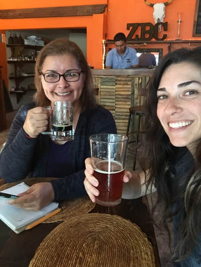 Two women smiling and enjoying drinks at a bar, one holding a glass of dark beer and the other a lighter amber beer. One has a notepad in front of her, while the background features a bartender and rustic decor, including a sign with the letters El Zopilote Brewery and Cocina offering craft beer and delicious cuisine.